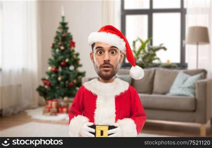 emotion, expression and winter holiday concept - surprised man in santa claus costume over christmas tree on home background (funny cartoon style character with big head). surprised man in santa costume over christmas tree