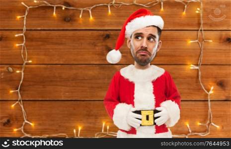 emotion, facial expressions and people concept - sad young man in costume of santa over garland lights on wooden background (funny cartoon style character with big head). sad santa over garland on wooden background