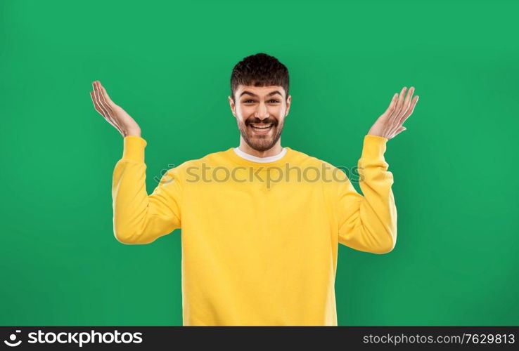 emotion, gesture and people concept - smiling young man in yellow sweatshirt shrugging over emerald green background. happy man in yellow sweater shrugging over green