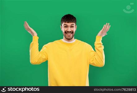 emotion, gesture and people concept - smiling young man in yellow sweatshirt shrugging over emerald green background. happy man in yellow sweater shrugging over green