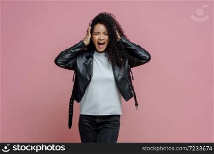Emotional crazy adult woman grabs head and shouts loudly, hears very loud music, keeps mouth opened, dressed in stylish leather jacket, isolated on pink background. People and emotions concept