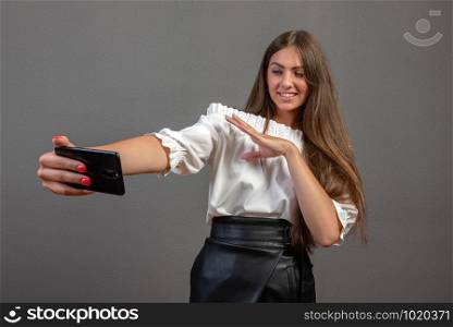 Emotional cute young woman posing isolated over gray wall background and take a selfie by mobile phone.