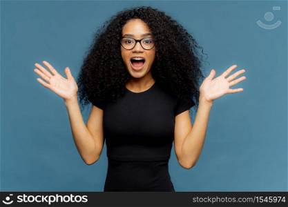 Emotional excited dark skinned lady raises palms, keeps mouth wide opened, exclaims loudly, wears black outfit, spreads hands, isolated on blue background. Impressed positive woman shouts with joy