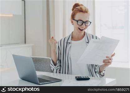 Emotional happy young ginger woman in eyewear holding paper with last mortgage payment banking notification, raising hand with clenched fist, reading document about tax refund from bank. Emotional happy young ginger woman holding paper with last mortgage payment banking notification