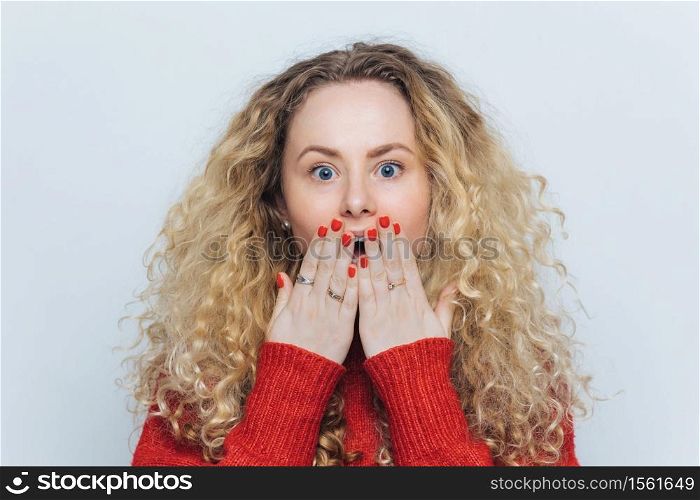 Emotional lovely woman with unexpected look covers mouth with hand, has red manicure, curly hair, isolated over white studio background. Shocked young cute female surprised to recieve present
