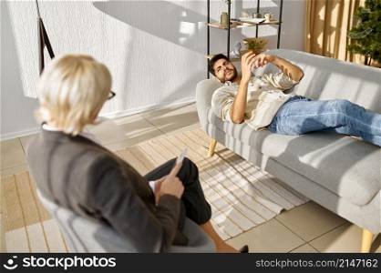 Emotional male patient lying on sofa explaining opinion to female psychologist during therapy session. Emotional young patient explaining opinion to psychologist