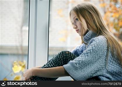 Emotional Portrait of a sad Girl with long blonde Hair. She is Sitting on the Window in the Rainy Autumn Day