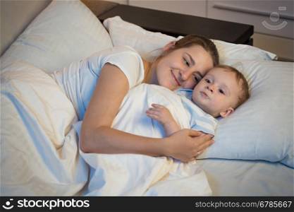 Emotional portrait of happy smiling mother embracing her baby in bed at night