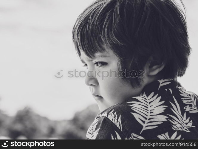 Emotional portrait of little boy lookig down with sad face, Side view head shot of kid with bored face standing alone in the park on sunny day, Child deep in though while looking at somr thing.