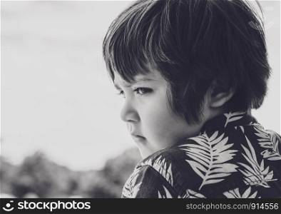 Emotional portrait of little boy lookig down with sad face, Side view head shot of kid with bored face standing alone in the park on sunny day, Child deep in though while looking at somr thing.