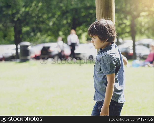 Emotional portrait of sad boy playing alone in playground with blurry car park background, Lonely Orphan child playing on his own, Dramatic portrait lost kid looking down with thinking face