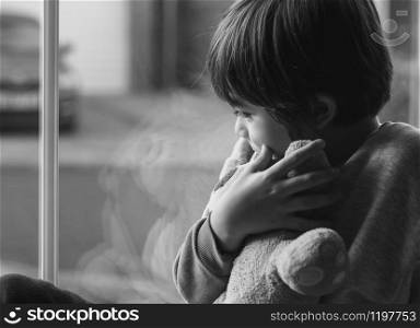 Emotional portrait of upset little boy sitting next to window, Kid sad face,Dramatic photo of Unhappy child looking down, Lonely boy sitting with his teddy bear, Spoiled children concept