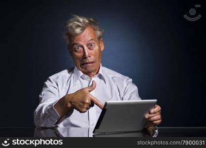 Emotional senior business man with an expressive face pointing to his laptop in horror and disbelief as he sits at a table against a dark studio background with copyspace