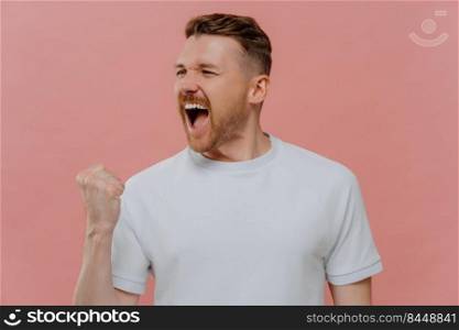 Emotional successful man shouts from joy clenches fist with triumph makes pump gesture cellebrates triumph dressed in casual white t shirt isolated over pink background. Body language concept