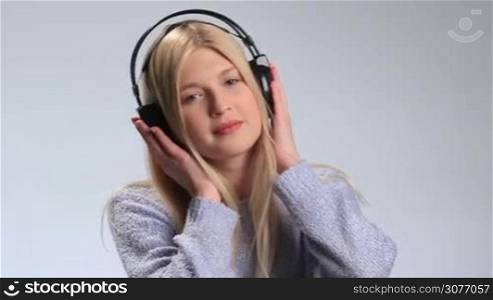 Emotional teenage girl enjoying music in headphones on white background. Girl swaying dreamily to music with her hands on headphones and closing her eyes. Smiling young woman blowing slow dreamy kiss at the camera.