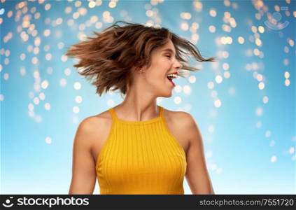 emotions and people concept - happy laughing young woman in mustard yellow top shaking head over holidays lights on blue background. happy young woman shaking head over lights