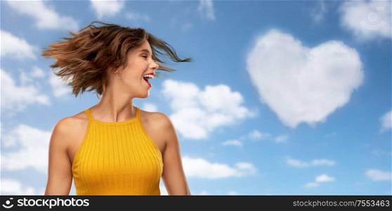 emotions and people concept - happy laughing young woman in mustard yellow top shaking head over blue sky and clouds background. happy young woman shaking head