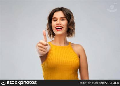 emotions and people concept - happy smiling young woman in mustard yellow top showing thumbs up over grey background. happy smiling young woman showing thumbs up