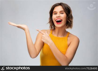 emotions and people concept - happy smiling young woman in mustard yellow top holding something on empty hand over grey background. happy young woman holding something on empty hand