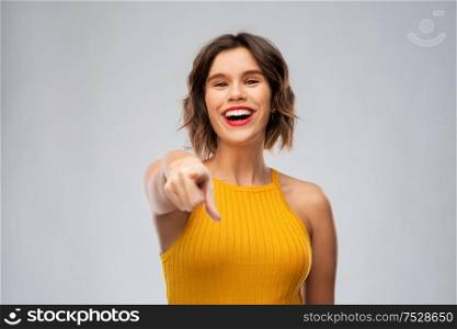 emotions and people concept - happy smiling young woman in mustard yellow top pointing to camera over grey background. happy smiling young woman pointing to camera