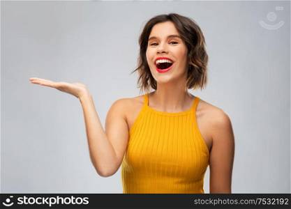 emotions and people concept - happy smiling young woman in mustard yellow top holding something on empty hand over grey background. happy young woman holding something on empty hand