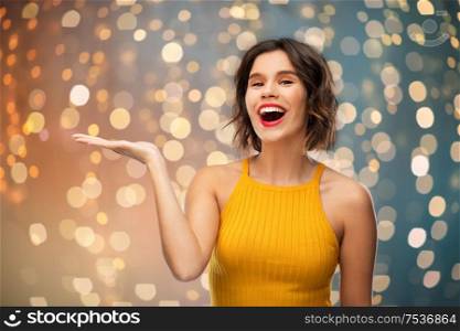 emotions and people concept - happy smiling young woman in yellow top holding something on empty hand over festive lights background. happy young woman holding something on empty hand