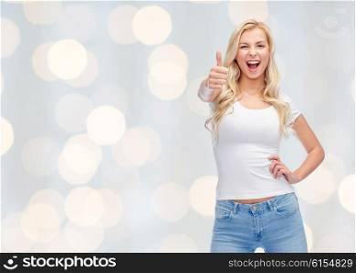 emotions, expressions, advertisement and people concept - happy smiling young woman or teenage girl in white t-shirt showing thumbs up over holidays lights background