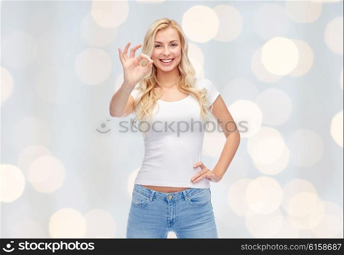 emotions, expressions, advertisement and people concept - happy smiling young woman or teenage girl in white t-shirt showing ok hand sign over holidays lights background