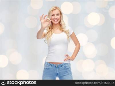 emotions, expressions, advertisement and people concept - happy smiling young woman or teenage girl in white t-shirt showing ok hand sign over holidays lights background