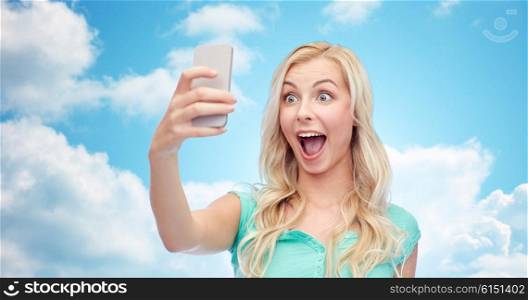 emotions, expressions and people concept - happy smiling young woman or teenage girl taking selfie with smartphone over blue sky and clouds background