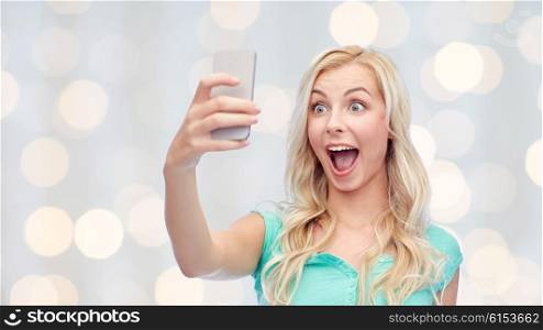 emotions, expressions and people concept - happy smiling young woman or teenage girl taking selfie with smartphone over holidays lights background
