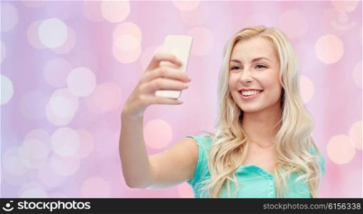 emotions, expressions and people concept - happy smiling young woman or teenage girl taking selfie with smartphone over pink holidays lights background