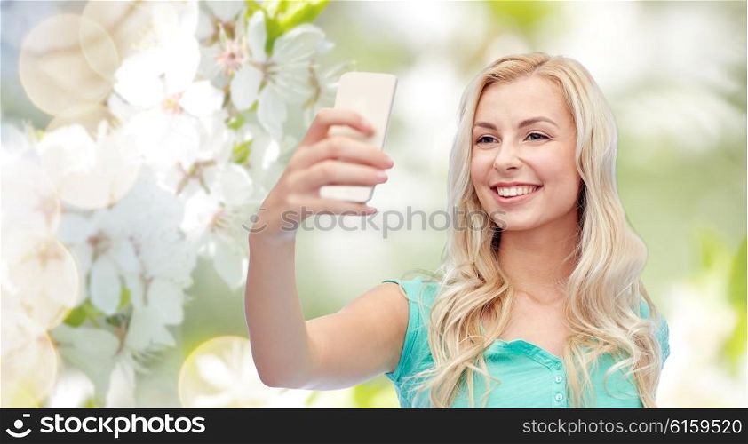 emotions, expressions and people concept - happy smiling young woman or teenage girl taking selfie with smartphone over natural spring cherry blossom background