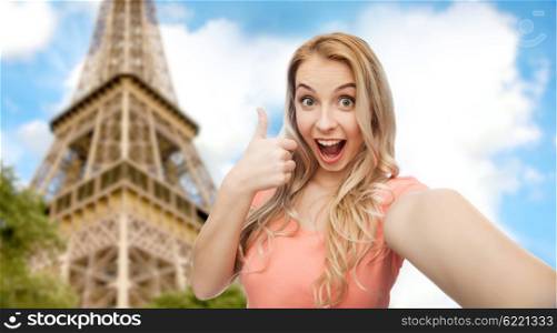 emotions, expressions and people concept - happy smiling young woman taking selfie and showing thumbs up over paris eiffel tower background