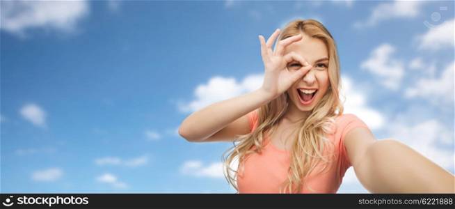 emotions, expressions and people concept - happy smiling young woman taking selfie over blue sky and clouds background