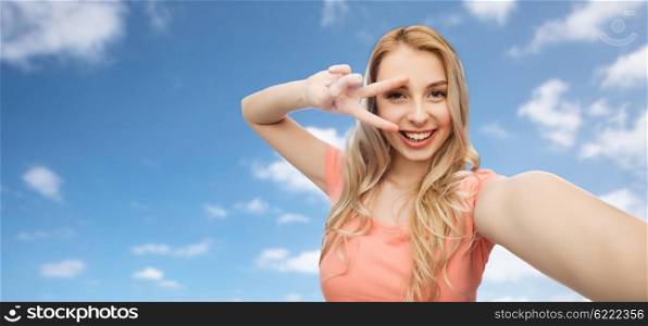 emotions, expressions and people concept - happy smiling young woman taking selfie and showing peace hand sign over blue sky and clouds background
