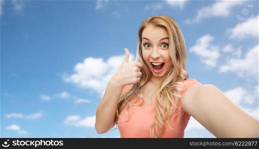 emotions, expressions and people concept - happy smiling young woman taking selfie and showing thumbs up over blue sky and clouds background