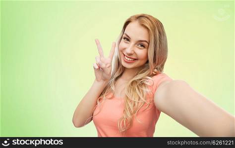 emotions, expressions and people concept - happy smiling young woman taking selfie and showing peace hand sign over green natural background