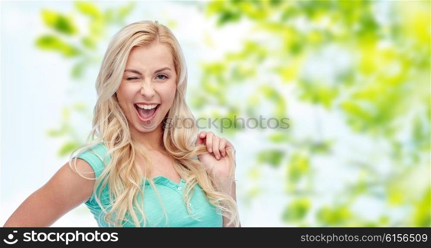 emotions, expressions, hairstyle and people concept - smiling young woman or teenage girl holding her strand of hair and winking over green natural background