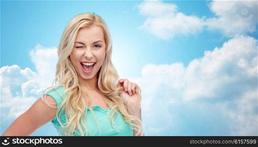 emotions, expressions, hairstyle and people concept - smiling young woman or teenage girl holding her strand of hair and winking over blue sky and clouds background