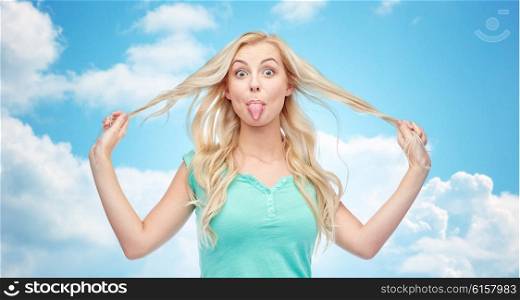 emotions, expressions, hairstyle and people concept - smiling young woman or teenage girl showing her tongue and holding strand of hair over blue sky and clouds background