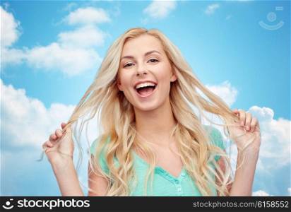 emotions, expressions, hairstyle and people concept - smiling young woman or teenage girl holding her strand of hair over blue sky and clouds background