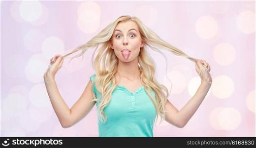 emotions, expressions, hairstyle and people concept - smiling young woman or teenage girl showing her tongue and holding strand of hair over pink holidays lights background