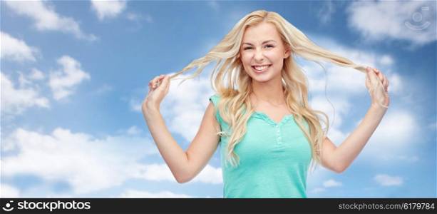 emotions, expressions, hairstyle and people concept - smiling young woman or teenage girl holding strands of her hair over blue sky and clouds background
