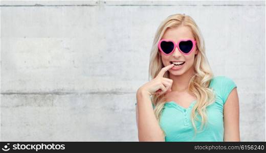 emotions, expressions, summer and people concept - smiling young woman or teenage girl in heart shape sunglasses over gray concrete wall background