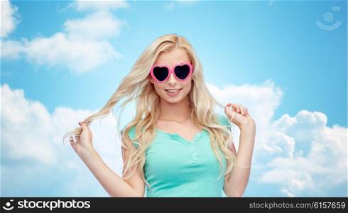 emotions, expressions, summer and people concept - smiling young woman or teenage girl in heart shape sunglasses holding her strand of hair over blue sky and clouds background
