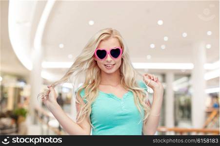 emotions, expressions, summer and people concept - smiling young woman or teenage girl in heart shape sunglasses holding her strand of hair over mall or shopping center background