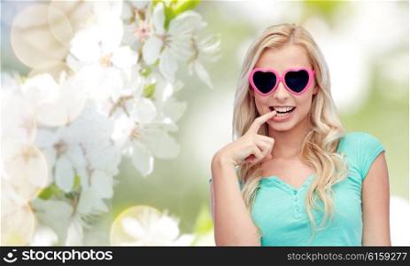 emotions, expressions, summer and people concept - smiling young woman or teenage girl in heart shape sunglasses over natural spring cherry blossom background