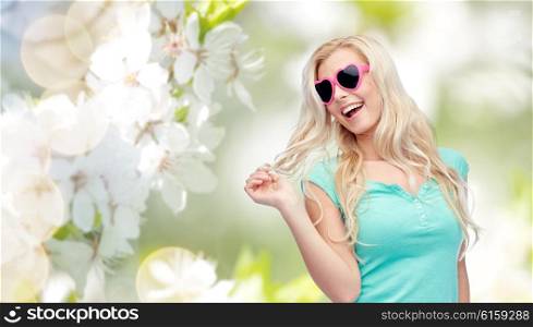 emotions, expressions, summer and people concept - smiling young woman or teenage girl in sunglasses holding her strand of hair over natural spring cherry blossom background