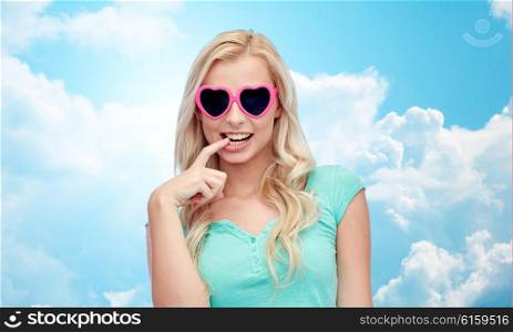 emotions, expressions, summer and people concept - smiling young woman or teenage girl in heart shape sunglasses over blue sky and clouds background